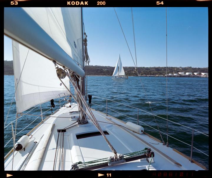A sailboat is on the water with another sailboat in the distance and a shoreline with buildings.