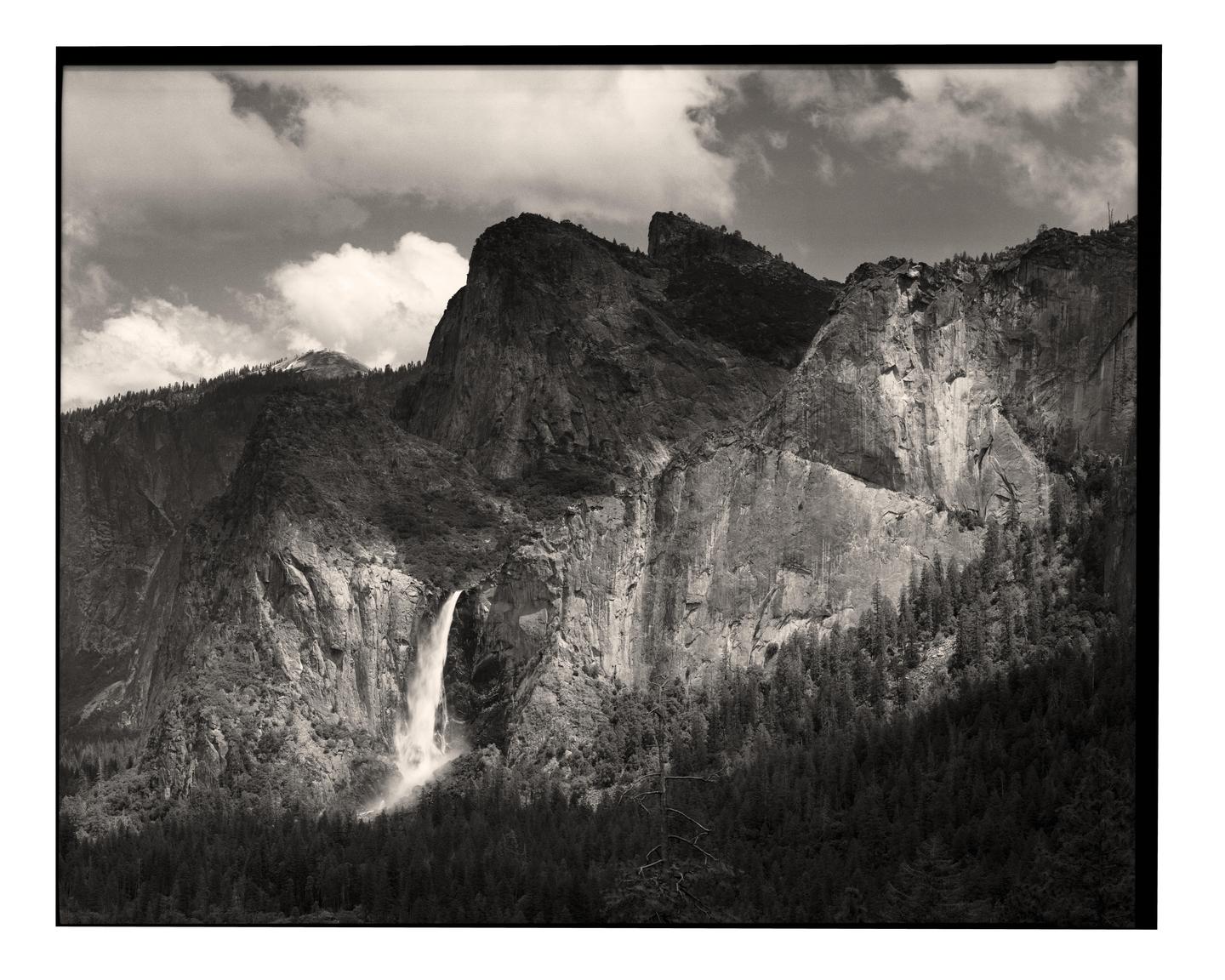 A waterfall cascades down a rocky mountain face with a forest at its base under a cloudy sky.