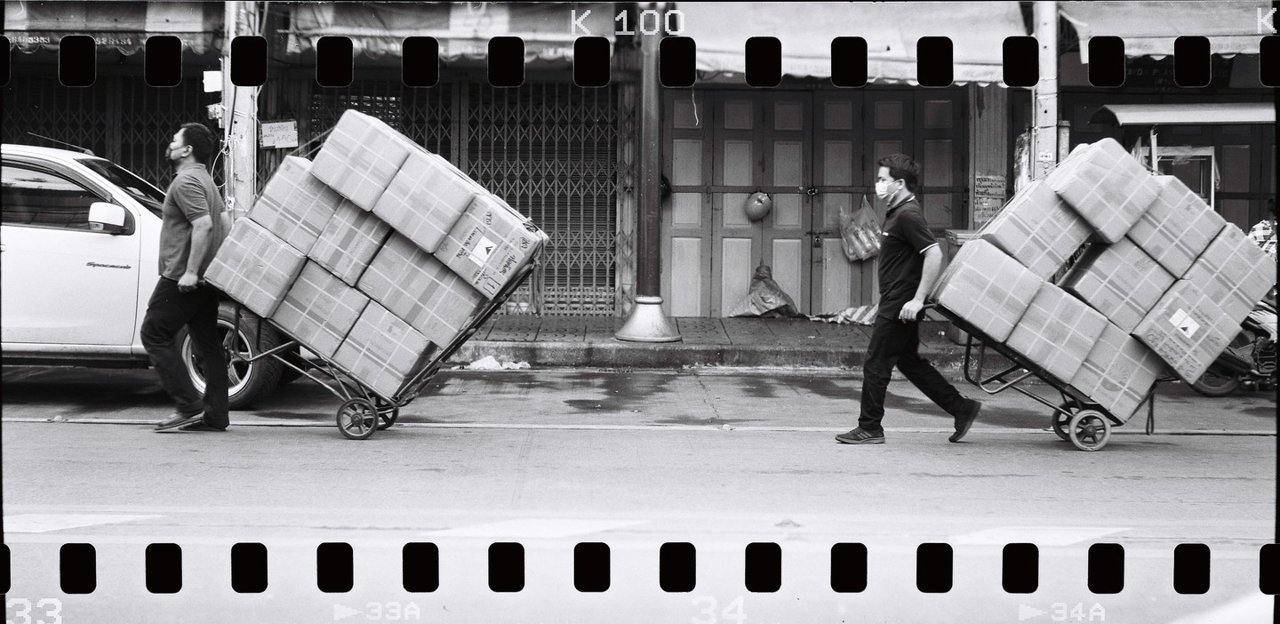 Two men wearing face masks push hand trucks stacked with boxes down a street past closed storefronts.