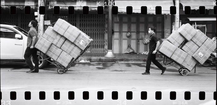 Two men wearing face masks push hand trucks stacked with boxes down a street past closed storefronts.