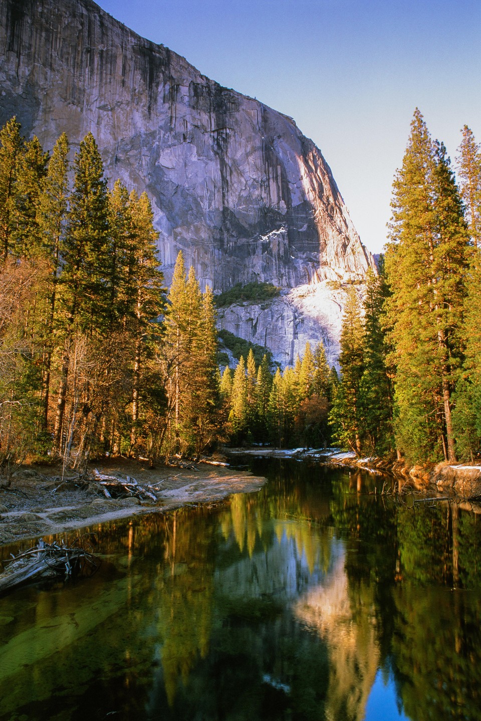 A river reflects a forest and a large rock face under a clear sky.