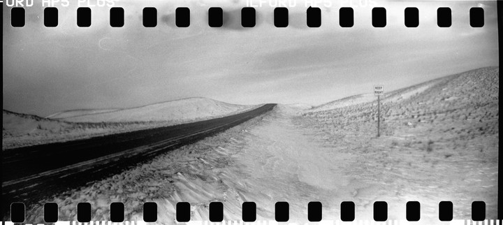 A long, empty road stretches into the distance, flanked by snow-covered hills and a "Keep Right" sign.