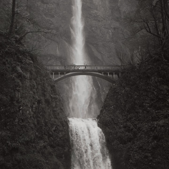 A bridge spans a tall waterfall with people standing on the bridge.