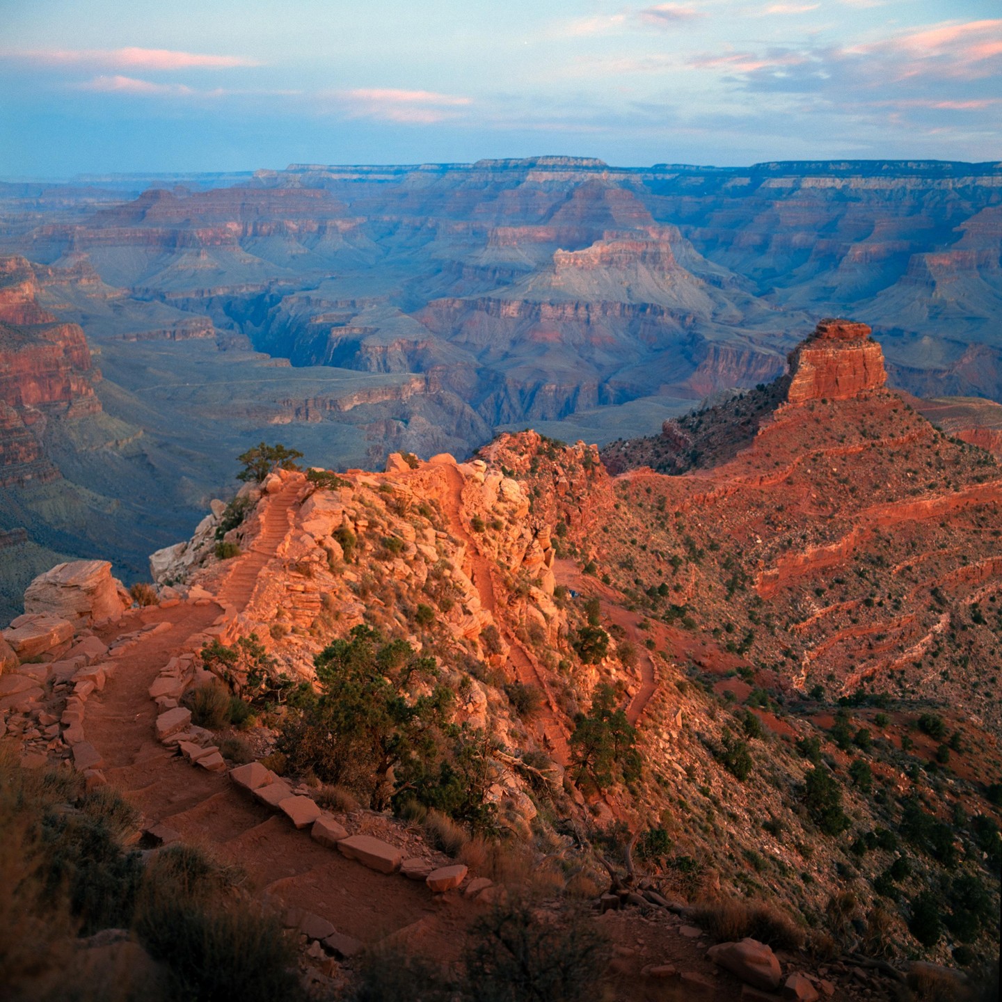 A winding trail leads down into the vast, layered landscape of the Grand Canyon at sunrise.