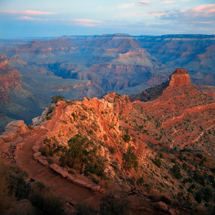 A winding trail leads down into the vast, layered landscape of the Grand Canyon at sunrise.