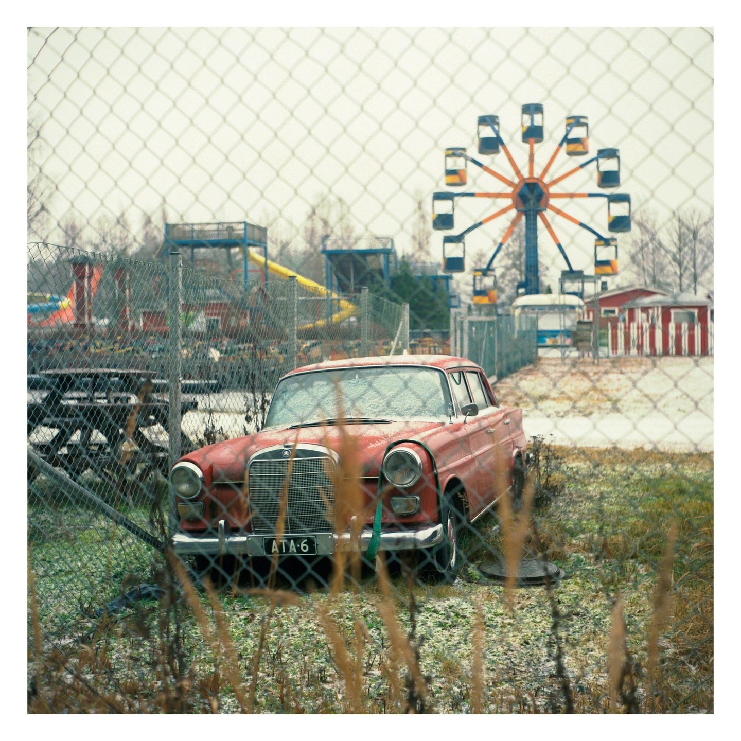 A red car is parked behind a chain link fence with an amusement park and ferris wheel in the background.