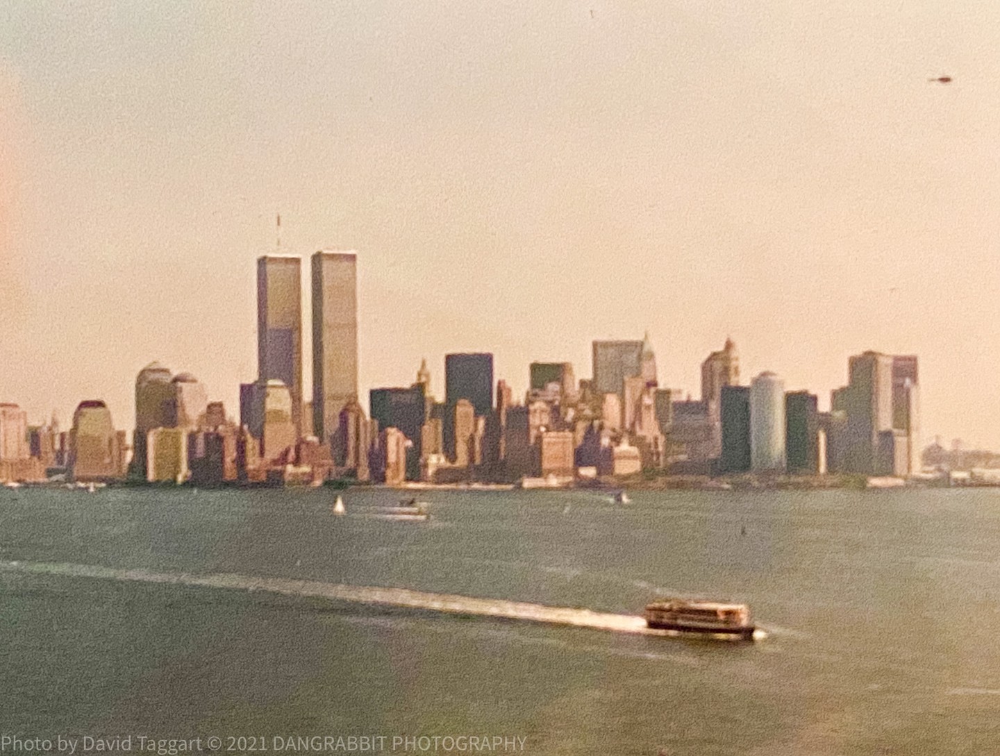 A ferry boat crosses a body of water with a city skyline and the twin towers in the background.