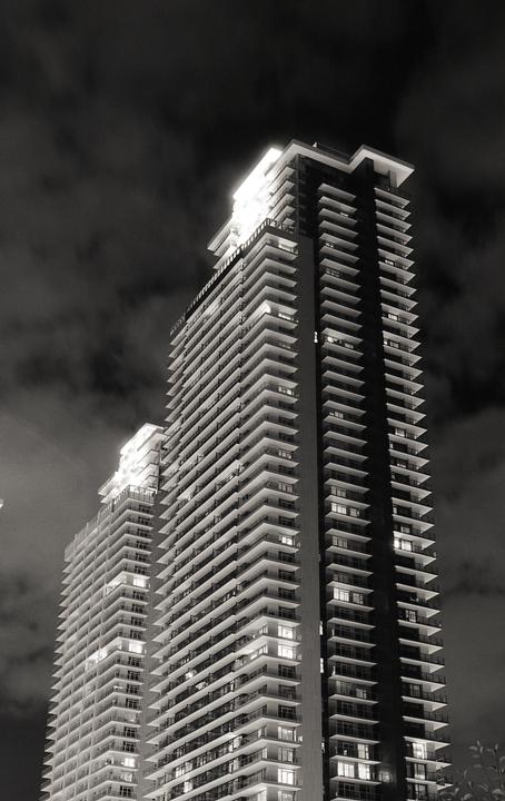 Two tall buildings with many balconies are illuminated at night against a cloudy sky.