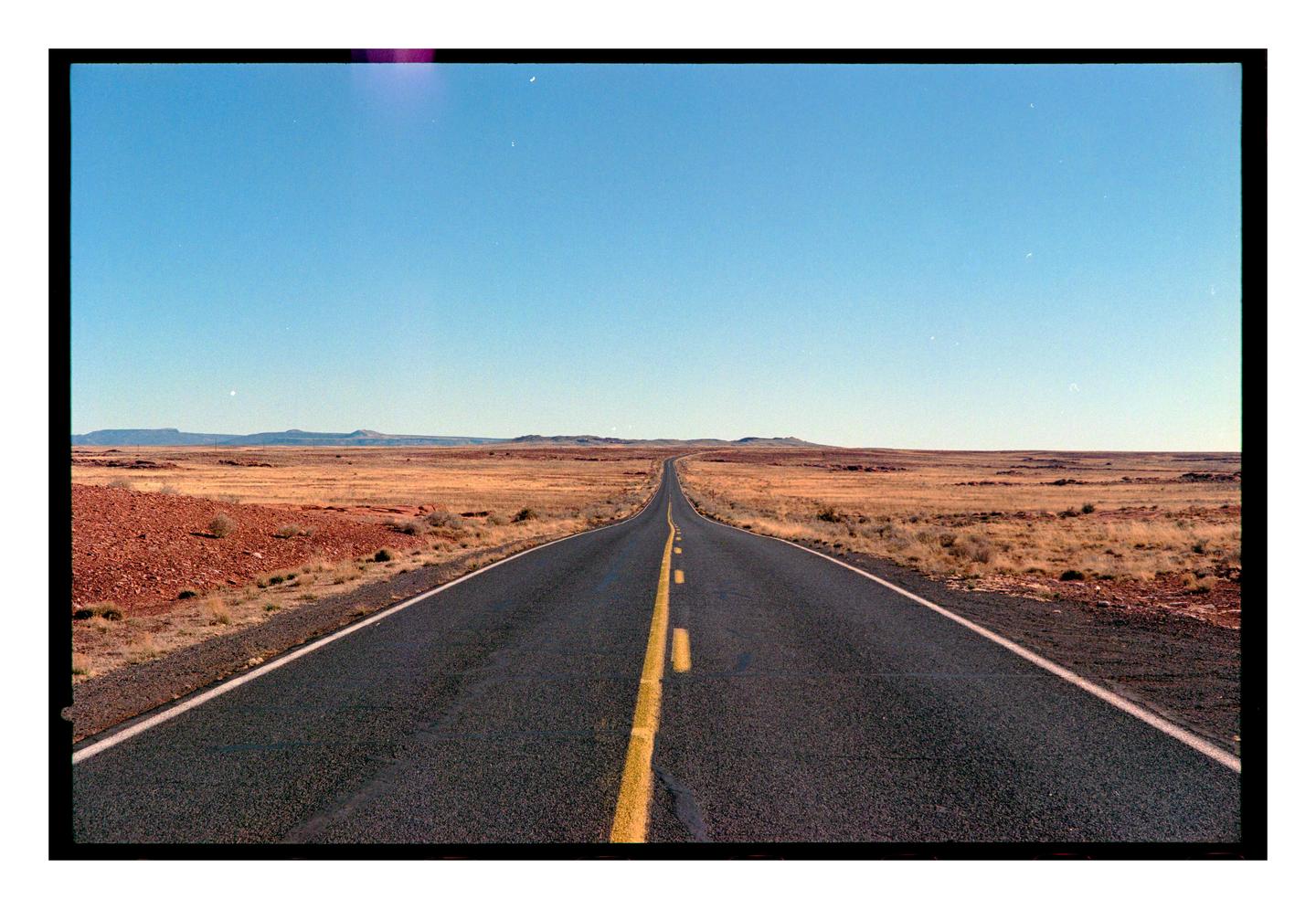A long, straight road stretches into the distance through a dry, open landscape under a clear sky.