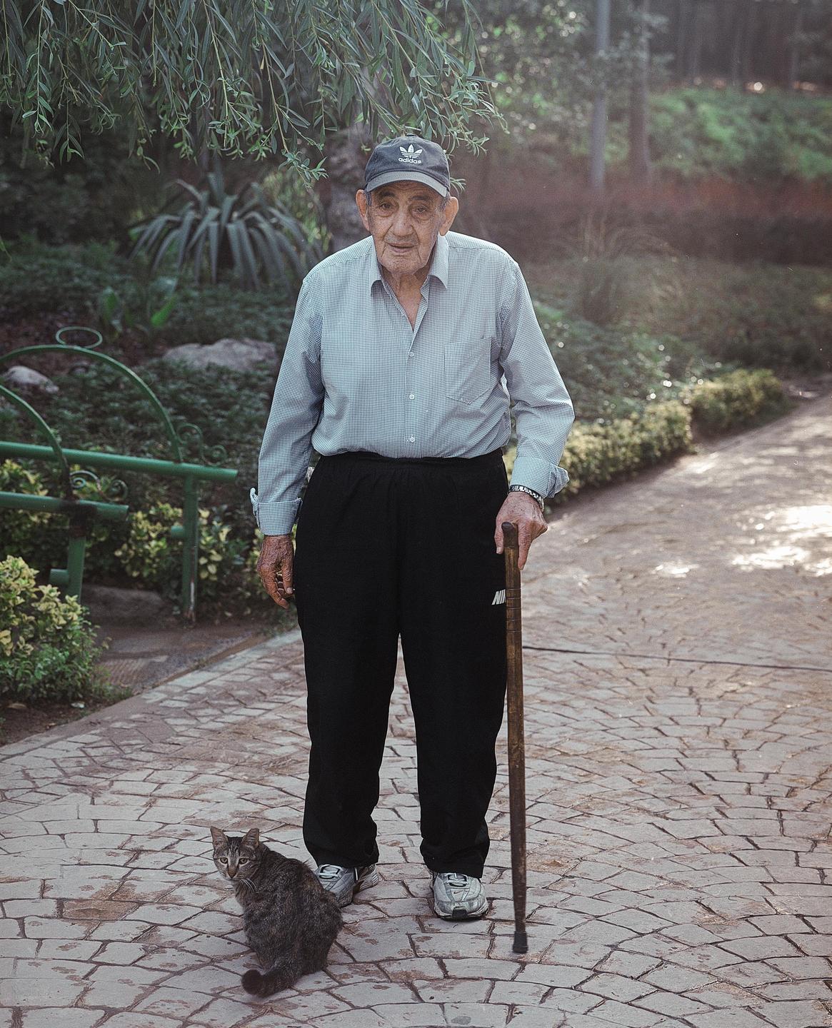 An elderly man with a walking stick stands on a paved path next to a cat.
