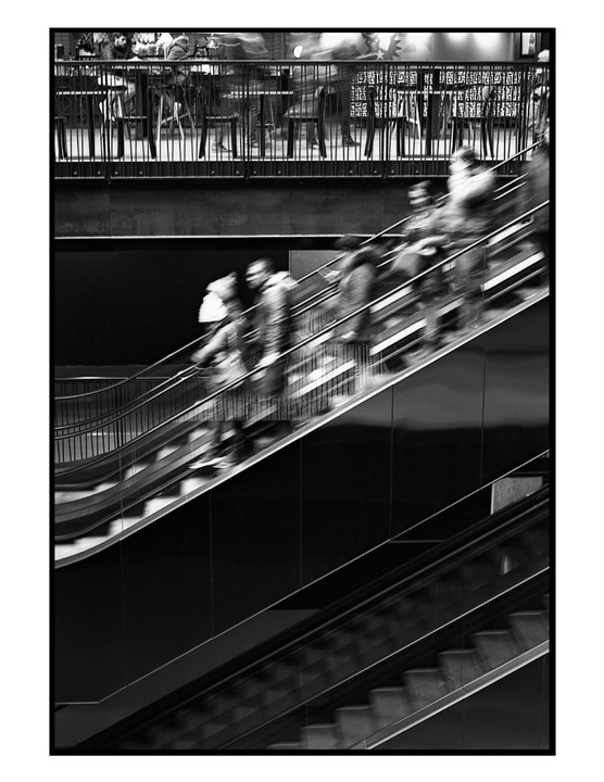 People ascend an escalator next to a cafe with blurred figures.