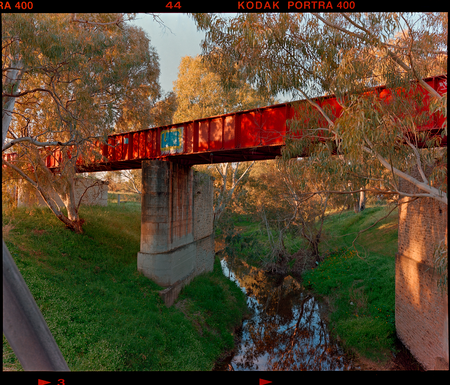 A red bridge crosses over a small stream surrounded by trees and green grass.