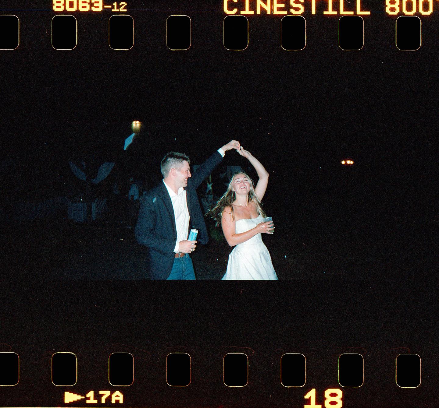 A couple dances at night, holding drinks and smiling.