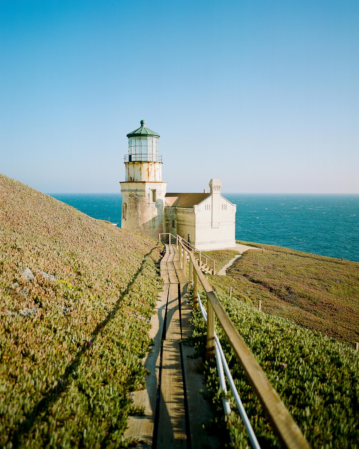 A wooden boardwalk leads to a white lighthouse on a grassy cliff overlooking the ocean.