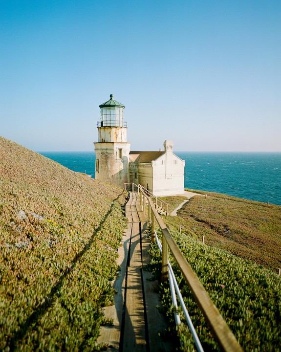 A wooden boardwalk leads to a white lighthouse on a grassy cliff overlooking the ocean.