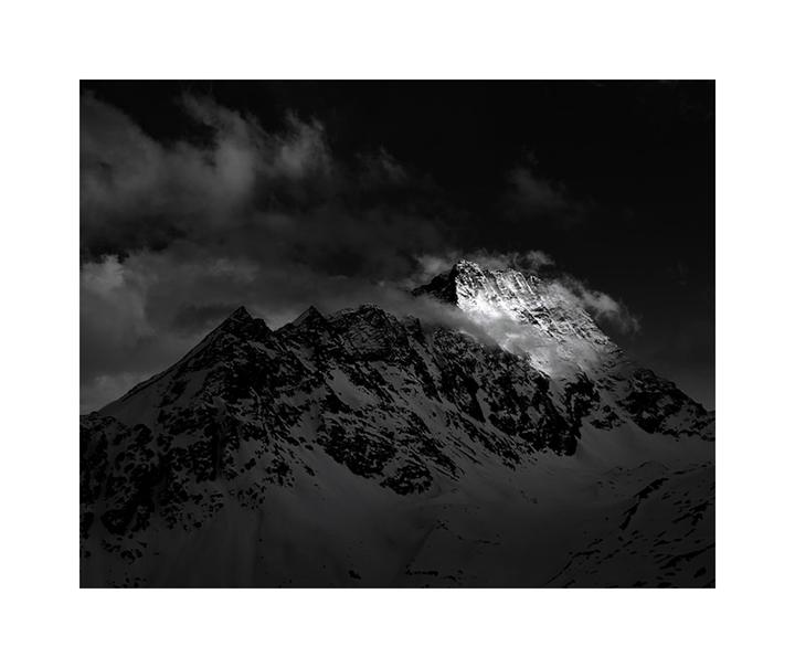 A snow-covered mountain peak is illuminated by a bright light against a dark, cloudy sky.