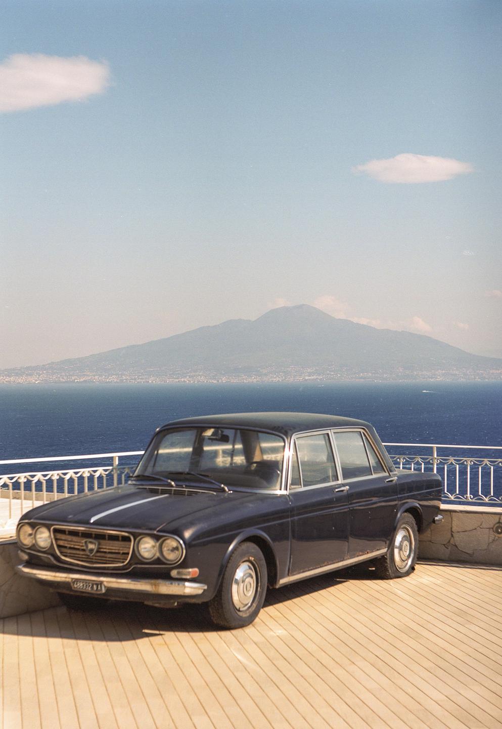 A dark blue vintage car is parked on a wooden deck overlooking a body of water with a mountain in the distance.