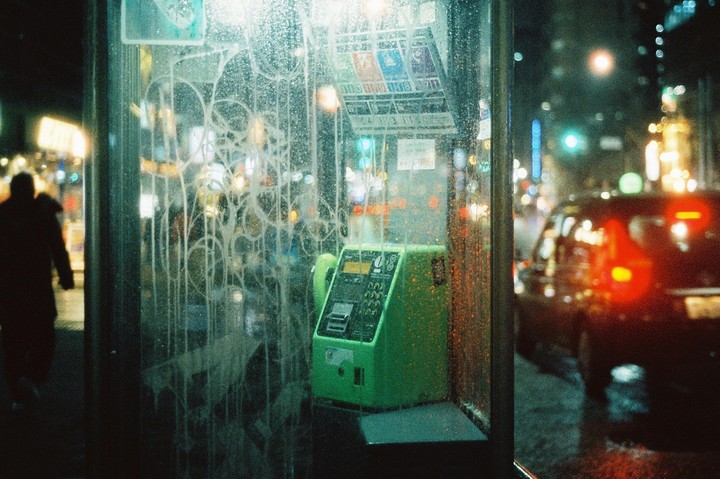 A green payphone is visible inside a phone booth on a rainy night street with a person walking by and a car in the background.