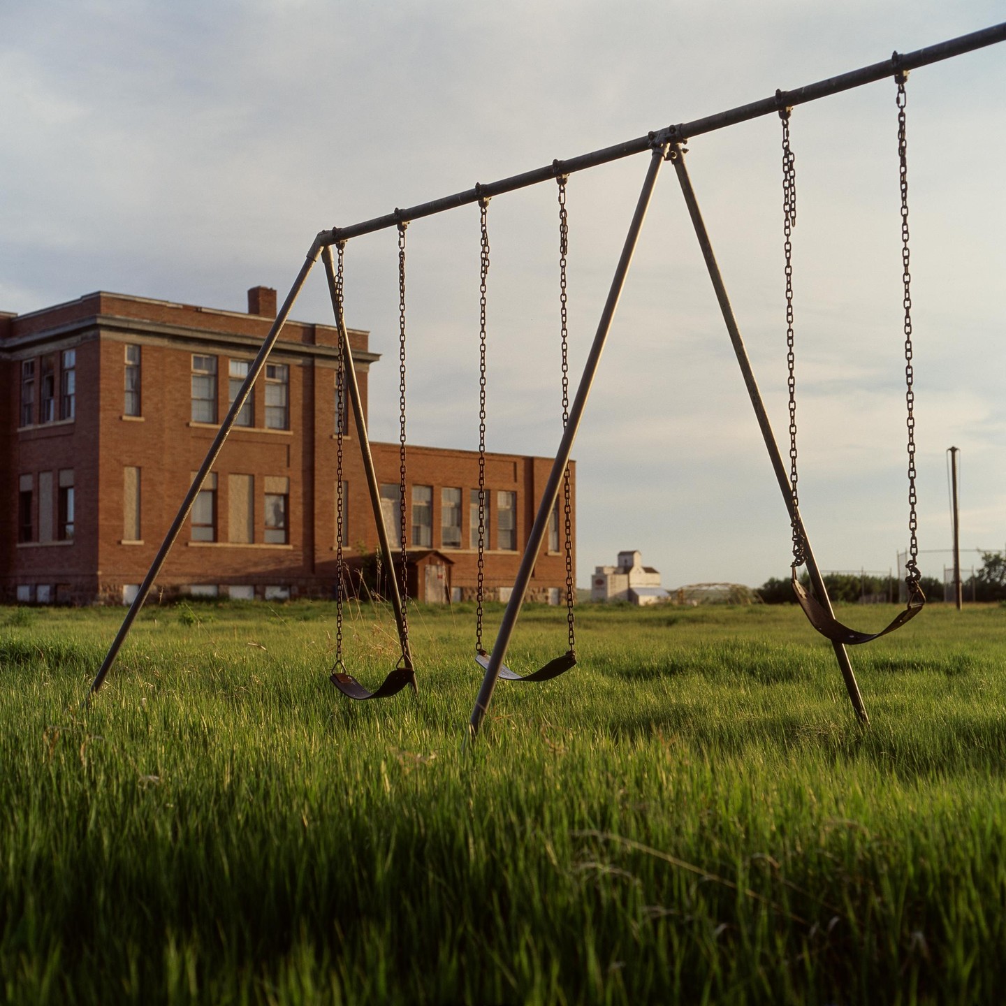 A swing set stands in tall grass in front of a brick building under a cloudy sky.