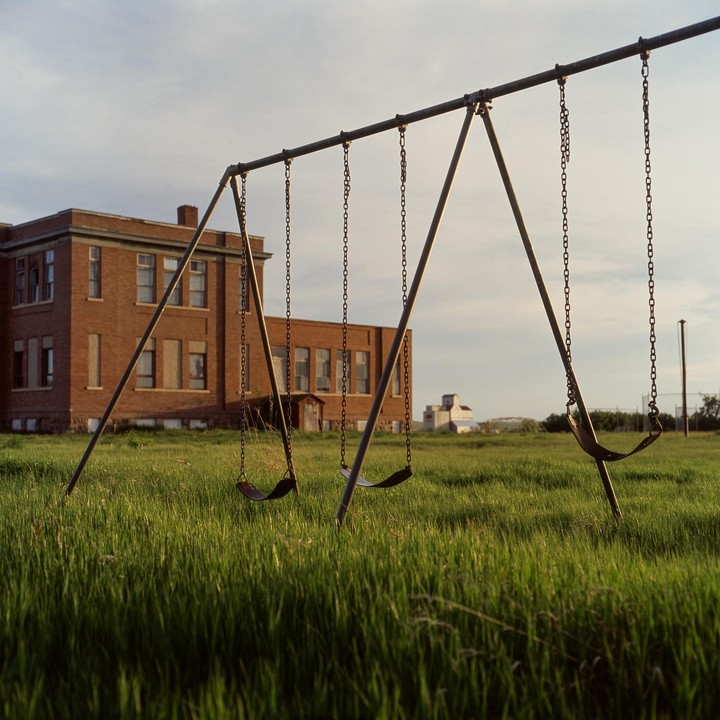 A swing set stands in tall grass in front of a brick building under a cloudy sky.