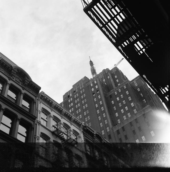 A low-angle view shows a tall building with a spire and a crane on its roof, flanked by smaller buildings and a fire escape, all under a bright sky.