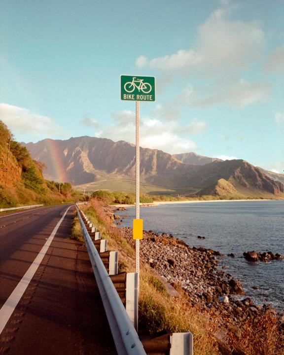 A bike route sign stands beside a coastal road with mountains and a rainbow in the distance.