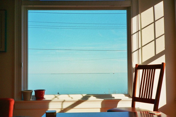 Sunlight streams through a window overlooking a vast expanse of blue water with power lines visible across the sky.