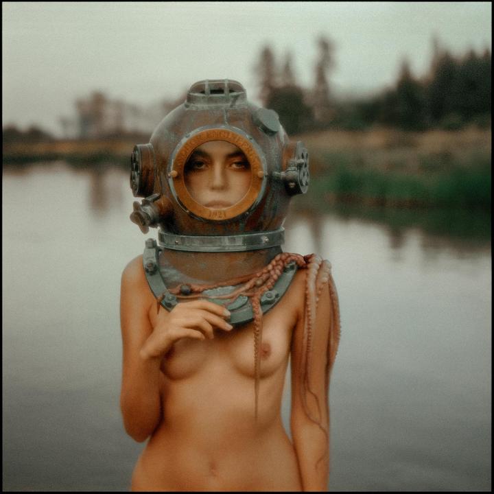 A woman wearing a diving helmet and an octopus around her neck stands in water.