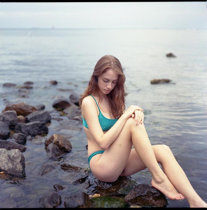 A woman in a bikini sits on rocks in the water, looking down at her lap.