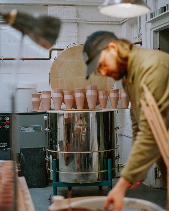 A man works in a pottery studio with several ceramic vessels on a shelf above a kiln.