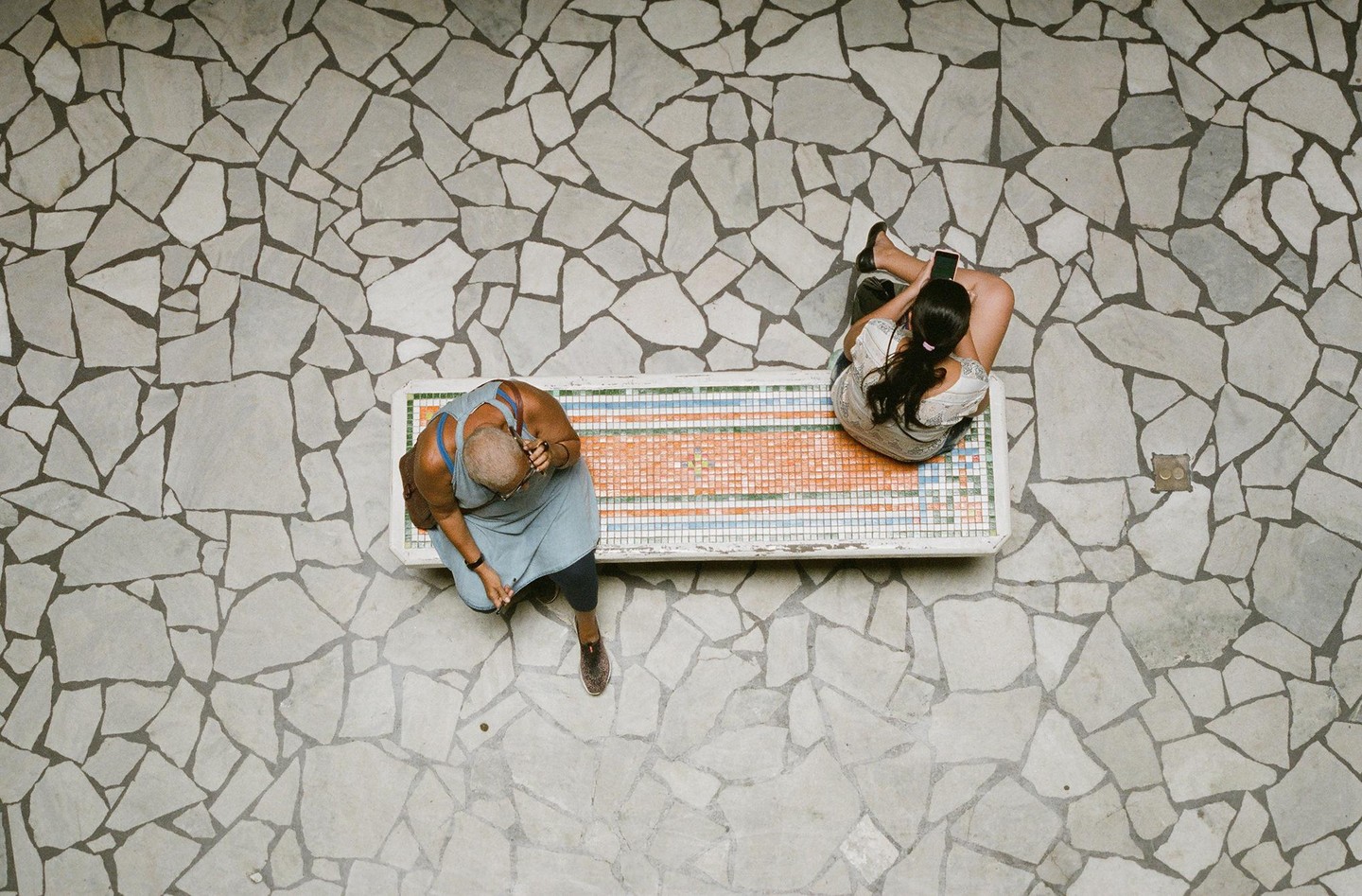 Two women sit on a tiled bench on a stone patio, viewed from above.