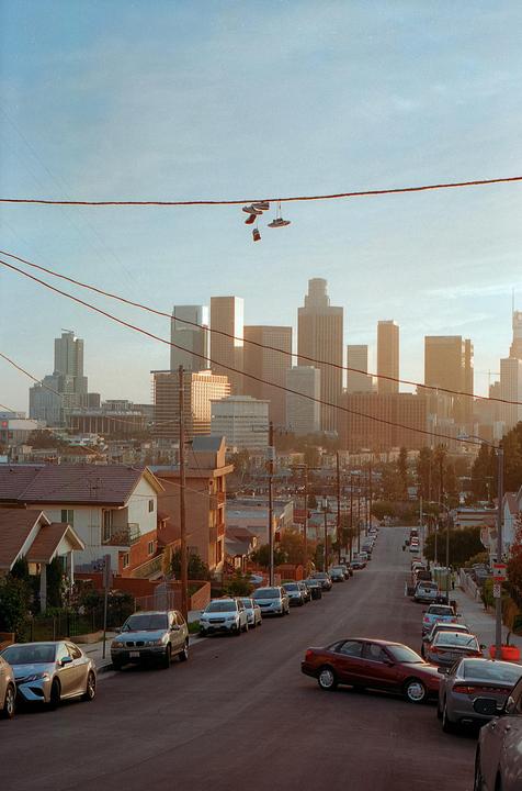 A pair of shoes hangs from a power line above a street with parked cars and a city skyline in the background.