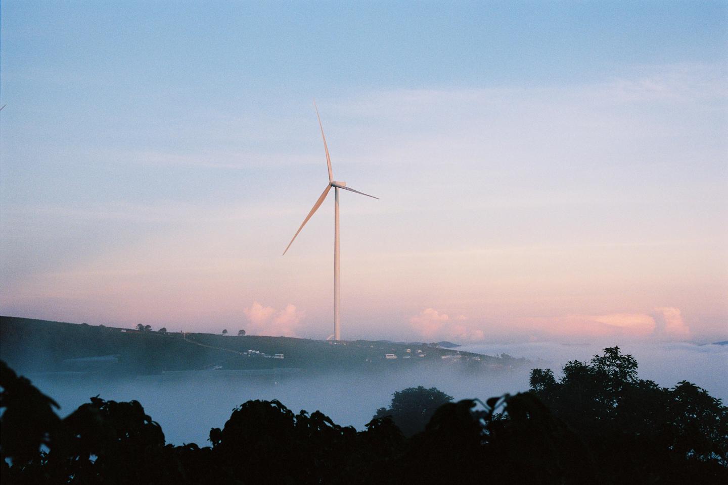 A wind turbine stands tall on a hill above a valley filled with fog at dusk.