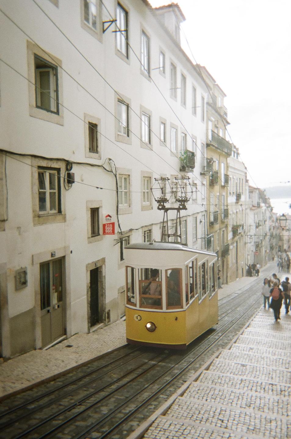 A yellow tram travels up a steep, narrow street lined with white buildings and people walking on the sidewalk.