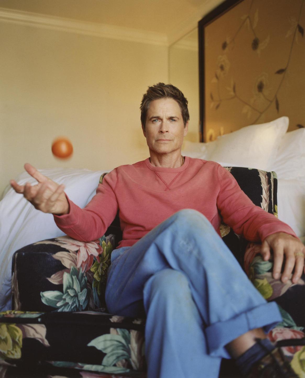 A man in a pink sweatshirt and blue pants sits in a floral armchair, tossing an orange in the air.