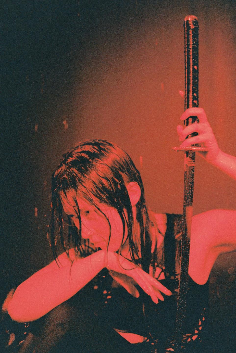 A woman with wet hair holds a katana in a rain studio.