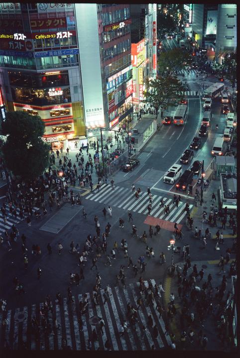 A high-angle view shows a busy street intersection with pedestrians and vehicles surrounded by buildings with illuminated signs at night.