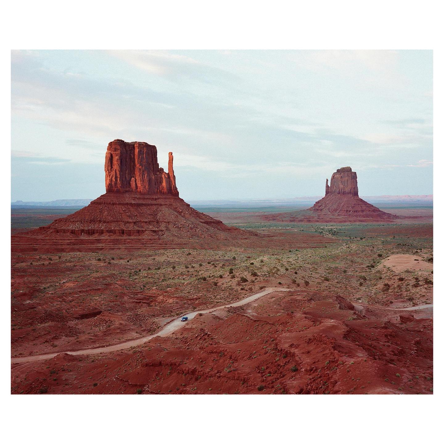 A small car drives on a dirt road through a desert landscape with two large rock formations under a pale sky.