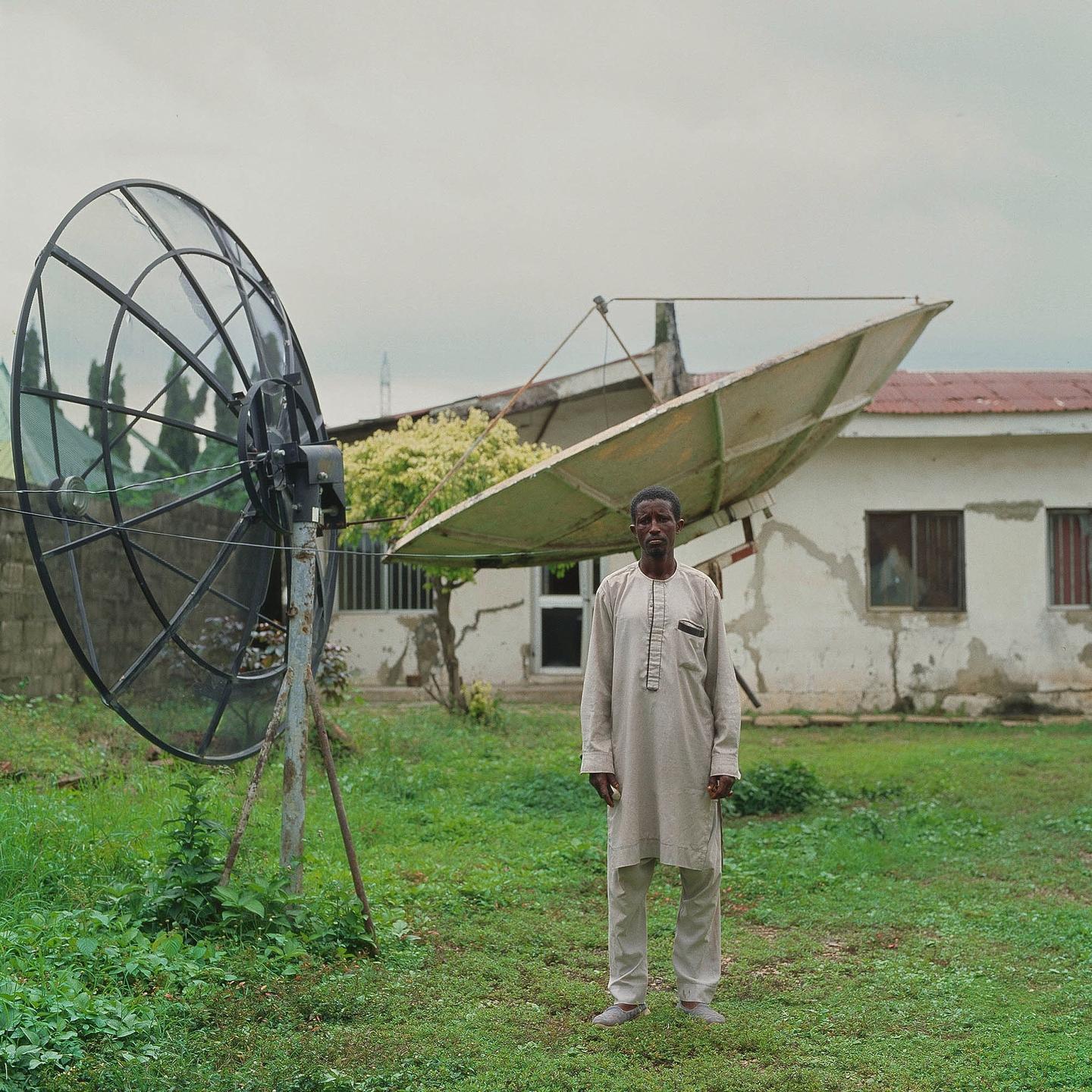 A man stands in a grassy yard in front of a building with two large satellite dishes.