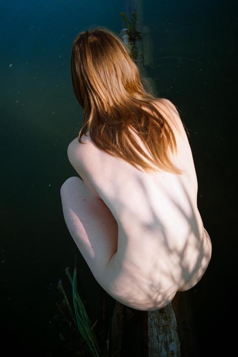 A woman with long red hair sits on a log over dark water, with shadows cast across her back.