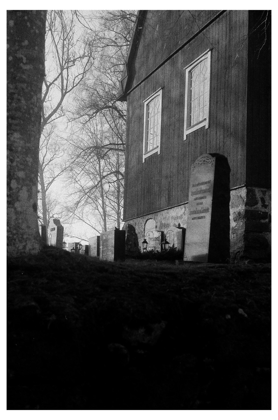 A black and white photograph shows a church building and gravestones on a hill, with bare trees in the background.