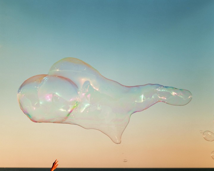 A large, iridescent soap bubble floats against a gradient sky with a hand reaching up from the bottom.
