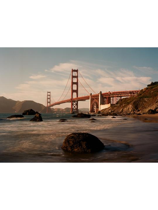 The Golden Gate Bridge spans a body of water with rocks in the foreground and hills in the background under a partly cloudy sky.