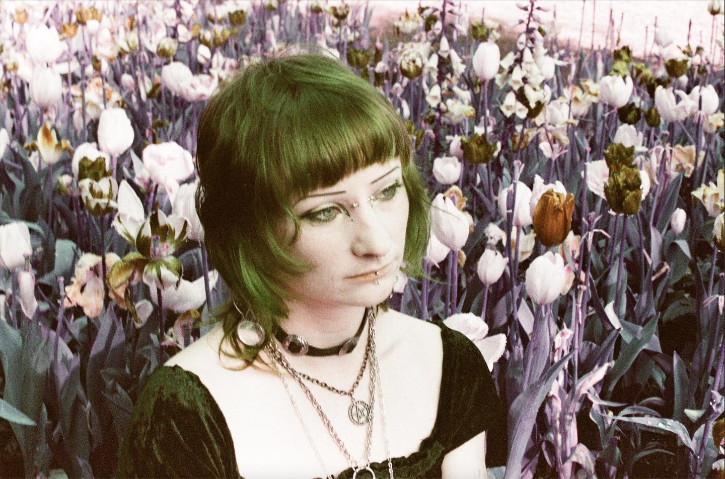 A woman with green hair and facial piercings is shown in a field of purple and white flowers.