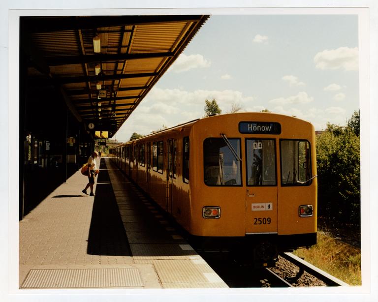 A yellow train is stopped at a station platform with a person walking in the shadow.