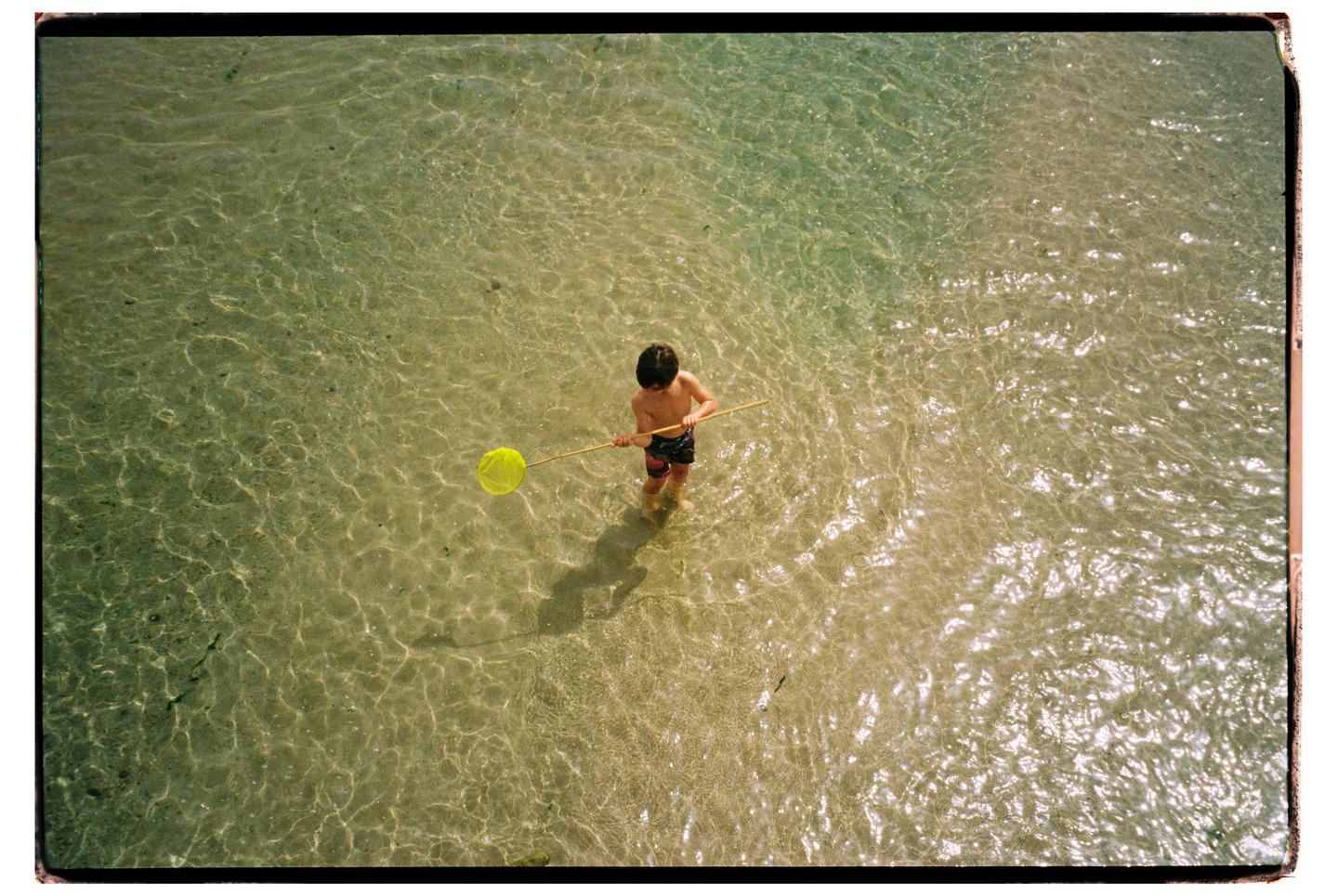 A boy stands in shallow, clear water holding a yellow net.