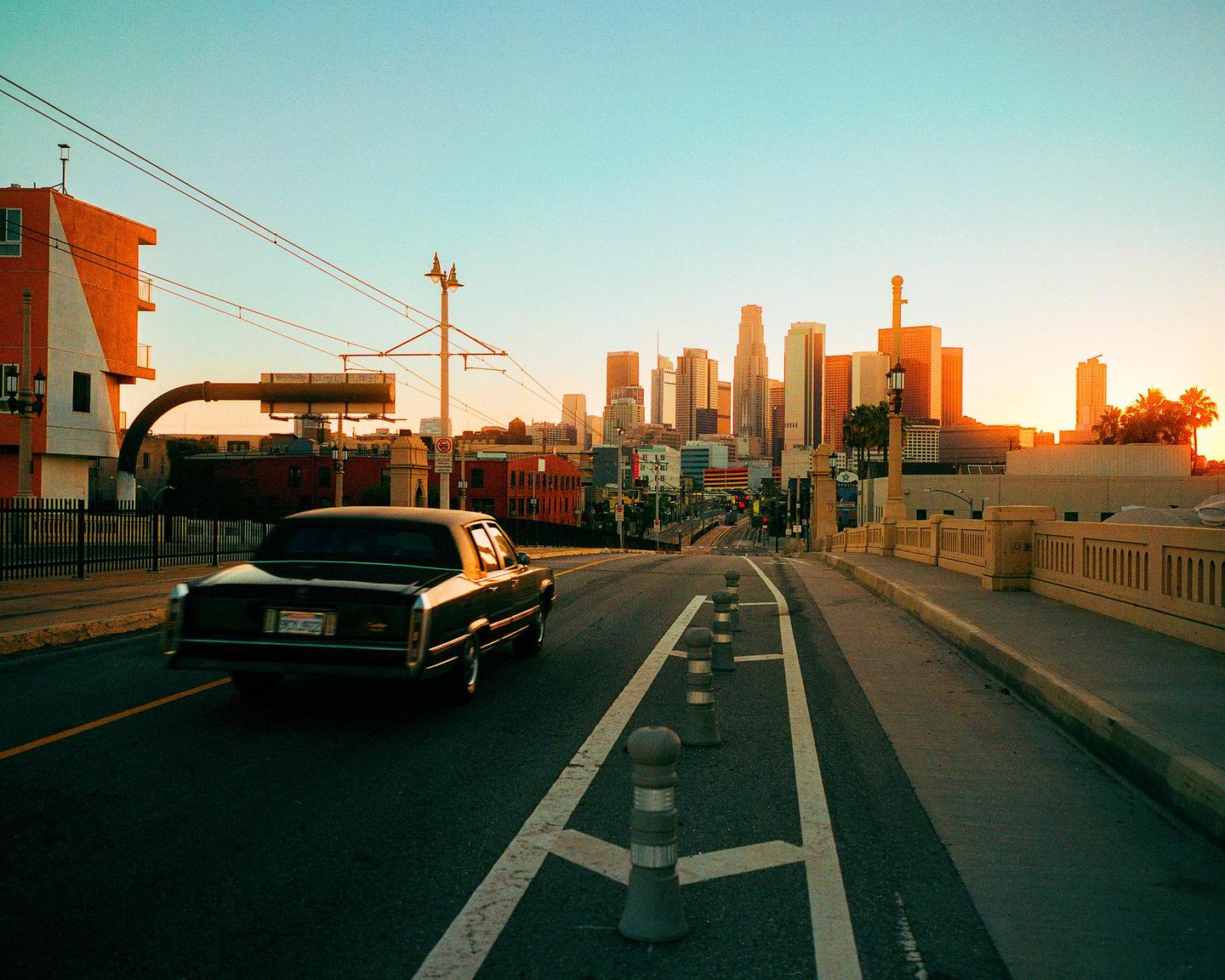 A black car drives on a street towards a city skyline at sunset.