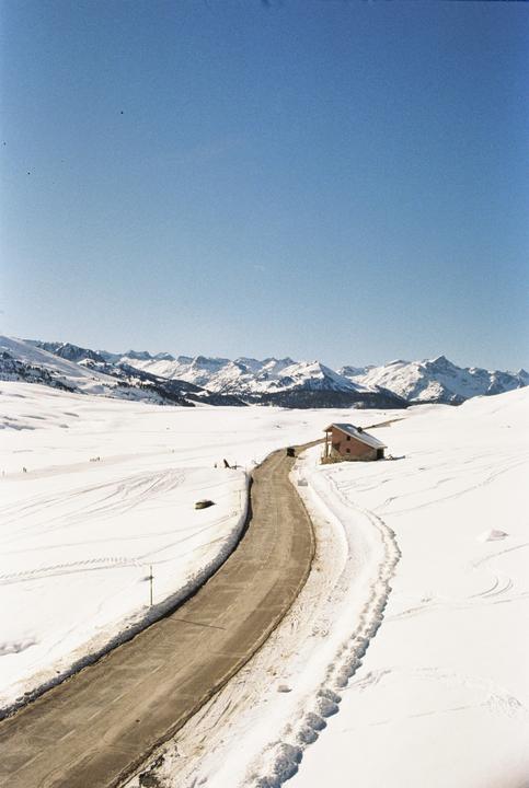 A winding road cuts through a vast snow-covered landscape with a small building and distant mountains under a clear sky.