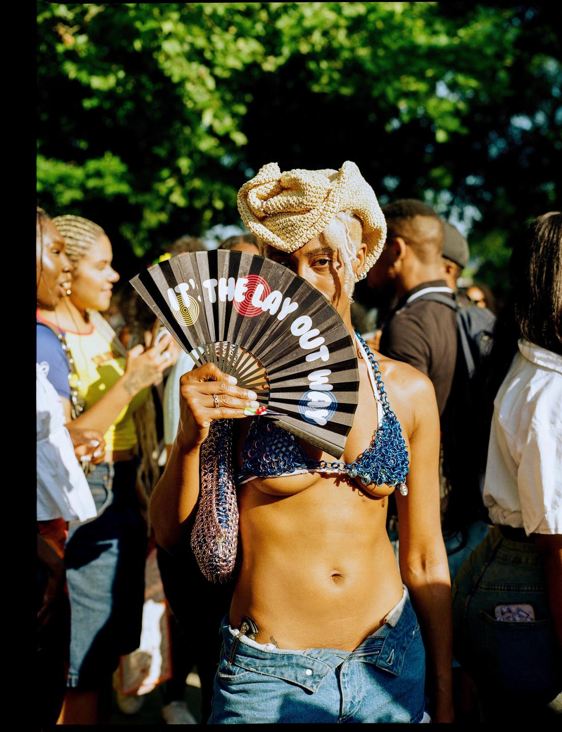 A woman in a crochet top and unbuttoned denim shorts holds a fan in front of her face, partially obscuring it.