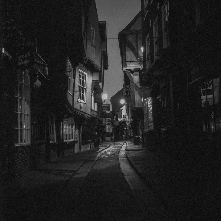 A narrow, winding street with historic buildings on both sides is illuminated by streetlights at night.